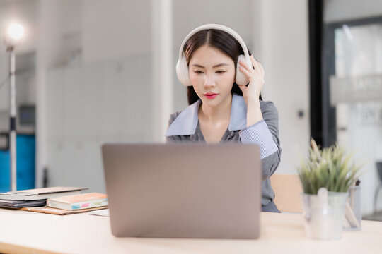 Asian woman concentrating at her desk, wearing white headphones while working on a laptop, embodying concepts of remote work, online learning, and focused productivity in a modern office environment