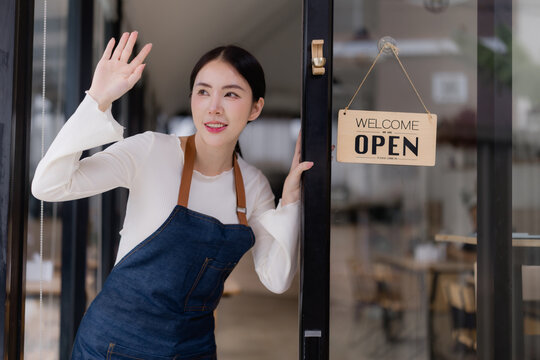 Young Asian woman wearing an apron standing in the doorway of her café or shop, waving hand, and smiling while opening a new small business, welcoming customers