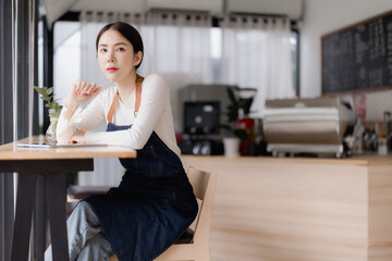 Young Asian woman wearing an apron and sitting at a wooden table inside a modern coffee shop, confidently planning for her business while looking at the camera