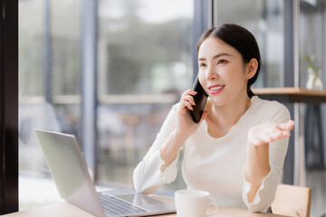 Asian executive woman smiling and talking on smartphone, working remotely using a laptop while sitting at a cafe table, illustrating modern communication and professional lifestyle