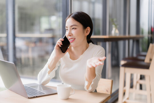 Asian woman confidently handling a phone call and laptop at a cafe table, smiling while managing remote work, freelancing and online business over coffee in a modern urban setting - Powered by Adobe