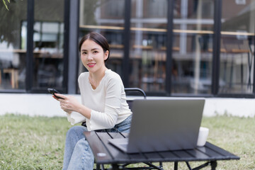 Asian woman using a smartphone and laptop outdoors in a relaxed setting, embracing remote work and digital nomad lifestyle with a coffee cup on the table