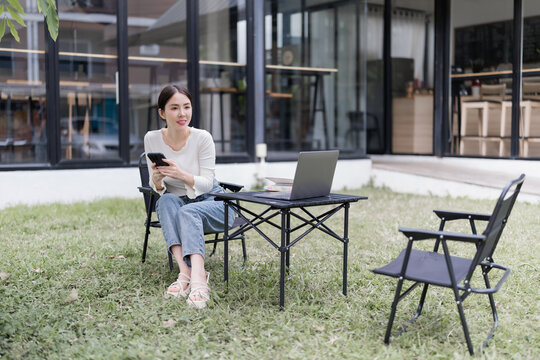 Young Asian woman sitting in a garden, holding a phone while looking away, with a laptop on a portable table, embodying the concept of flexible remote work and digital nomad lifestyle - Powered by Adobe