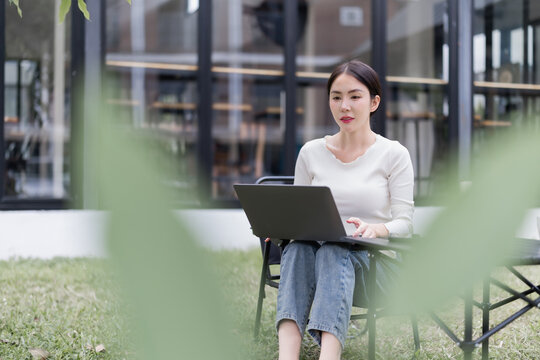 Asian woman concentrating while typing on a laptop, enjoying remote work in a casual outdoor setting on a sunny day in a cafe garden, embracing freedom and flexibility - Powered by Adobe