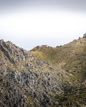 View of jagged, rocky mountains rise against a soft, overcast sky, a solitary structure perched atop a distant peak, Mallorca, Balearic Islands, Spain.