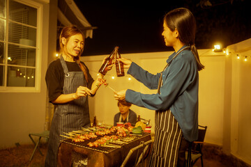 Two Asian women in striped aprons toast with glasses or beer bottles during a backyard night BBQ. Skewered marinated meats grill densely, creating a warm,festive atmosphere of friendship celebration