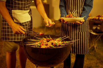 An Asian woman enjoys a cozy backyard BBQ, preparing or serving skewered meats and vegetables. Warm golden lights create a festive, relaxed atmosphere, highlighting friendship, celebration, outdoor