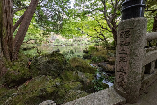View of mossy stones and lush greenery reflecting in tranquil waters under the canopy of trees, with a stone pillar in the foreground, Narita, Chiba, Japan.