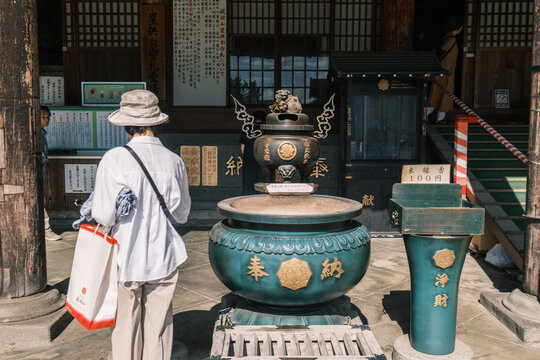 View of a woman in a hat and white shirt stands before a large, ornate bronze incense burner at a temple, bathed in soft light, Narita, Chiba, Japan.