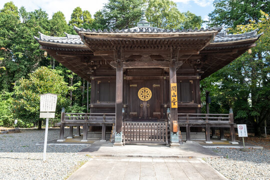 View of an old wooden temple standing peacefully among lush trees, with intricate carvings and a golden emblem, reflecting serenity and heritage, Narita, Chiba, Japan.