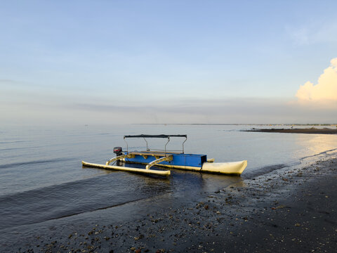 View of a blue and white outrigger boat resting on a dark sandy beach under a serene sky where the sea meets the horizon, Banyuwangi, East Java, Indonesia.