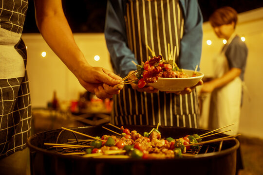 An Asian woman enjoys a cozy backyard BBQ, preparing or serving skewered meats and vegetables. Warm golden lights create a festive, relaxed atmosphere, highlighting friendship, celebration, outdoor
