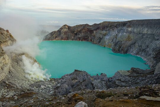 View of a vibrant turquoise lake nestled within a rugged volcanic crater, wisps of smoke rising against the textured rock formations, Banyuwangi, East Java, Indonesia.