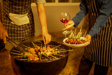An Asian woman enjoys a cozy backyard BBQ, preparing or serving skewered meats and vegetables. Warm golden lights create a festive, relaxed atmosphere, highlighting friendship, celebration, outdoor