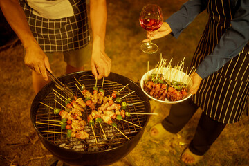 An Asian woman enjoys a cozy backyard BBQ, preparing or serving skewered meats and vegetables. Warm golden lights create a festive, relaxed atmosphere, highlighting friendship, celebration, outdoor