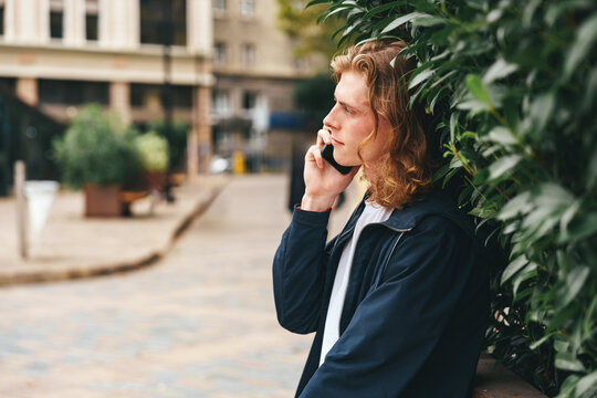 Young man talking on phone while leaning against greenery in a city setting