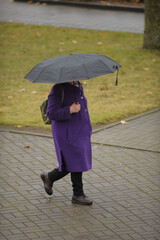 A middle-aged woman in a lilac coat walks down the street with an umbrella in rainy weather