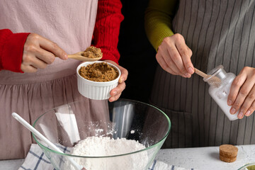 Two people preparing baking ingredients—adding brown sugar and salt to flour in a glass bowl. Holiday cookie baking scene in a cozy kitchen
