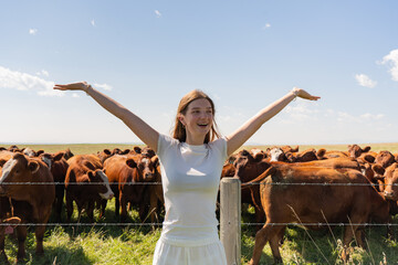 Girl with arms raised among curious cattle in a sunny pasture setting. © Viks