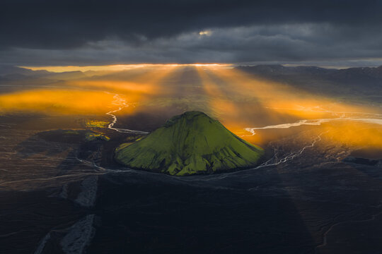 Aerial view of golden light piercing through dark clouds illuminating a green mountain surrounded by glacial rivers, Highlands Of Iceland, Sveitarf&eacute;lagio Hornafjorour, Iceland.