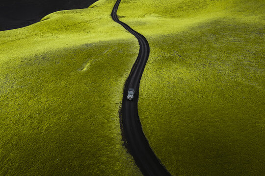 Aerial view of a lone car winding through a vibrant green landscape cut by a stark dark road under a brooding sky, Highlands Of Iceland, Sveitarf&eacute;lagio Hornafjorour, Iceland.