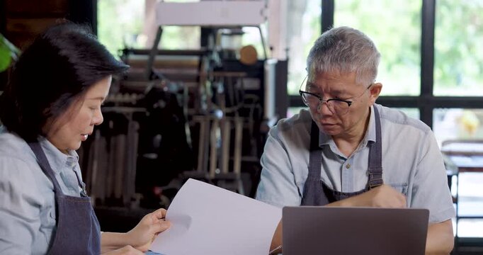 Asian elderly female and male cafe owners in aprons reviewing business accounting report together at cafe table, teamwork communication highlighting cooperation partnership and financial management
