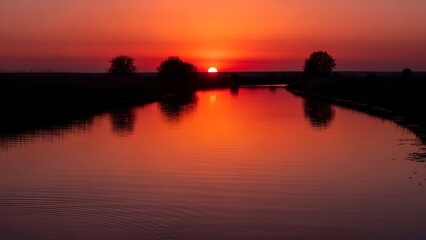 Obraz premium Crimson sunset reflects on calm river, silhouettes trees and distant fields. A moment of peace
