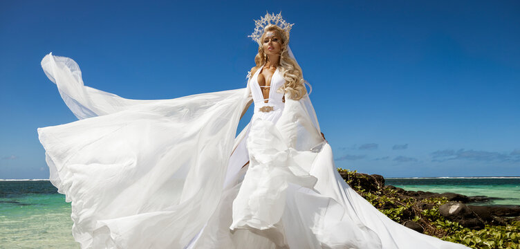 Elegant woman in flowing white dress and crown posing by turquoise ocean on Mauritius beach under blue sky, luxury fashion photoshoot with wind and movement