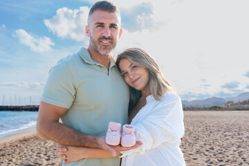 Expecting couple standing on sandy beach, smiling at camera, pregnant woman holding tiny pink baby shoes with love and tenderness. Bright sky, calm sea and sunlight create peaceful,emotional maternity