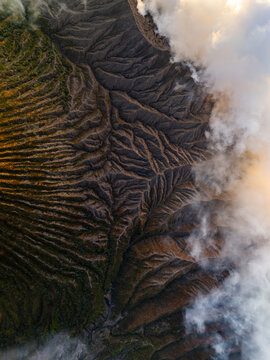 Aerial view of the rugged, textured landscape of Mount Bromo, where earthy tones meet the soft embrace of swirling clouds, East Java, Indonesia.
