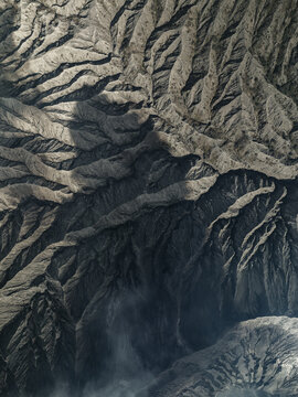 Aerial view of a stark, rugged landscape with deep shadows contrasting sharply with the sunlit peaks of Mount Bromo, East Java, Indonesia.