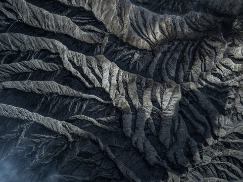Aerial view of rugged mountain terrain carved by deep ravines, displaying stark contrasts of light and shadow across the landscape, Mount Bromo, East Java, Indonesia.