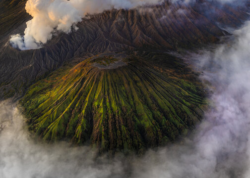 Aerial view of the dramatic volcanic landscape where verdant slopes meet the stark, smoky crater, framed by ethereal clouds, Mount Bromo, East Java, Indonesia.