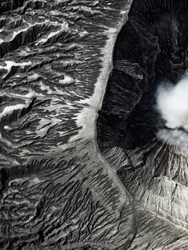 Aerial view of the dark, brooding caldera of Mount Bromo, where smoke plumes rise against the stark, rugged landscape, Mount Bromo, East Java, Indonesia.
