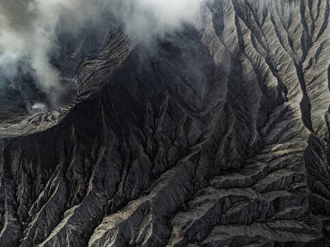 Aerial view of rugged, volcanic terrain etched with deep ravines and shrouded in ethereal smoke, creating a stark contrast of light and shadow, Mount Bromo, East Java, Indonesia.
