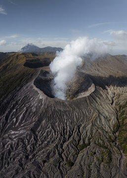 Aerial view of a smoking volcano crater amidst a rugged, textured landscape under a clear sky, Mount Bromo, East Java, Indonesia.