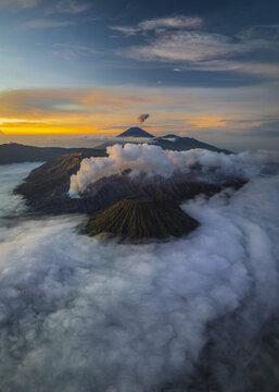 Aerial view of Mount Bromo piercing through a sea of swirling clouds under a vibrant dawn sky, Mount Bromo, East Java, Indonesia.