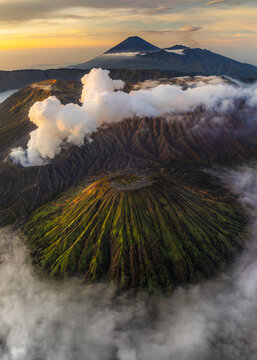 Aerial view of the majestic Mount Bromo volcano, its peak shrouded in ethereal clouds, a stark contrast to the lush green slopes below, Mount Bromo, East Java, Indonesia.