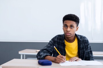Teen student sitting at desk in classroom, focused on writing test or assignment. Back to school, education and exam preparation concept. Horizontal indoors shot.