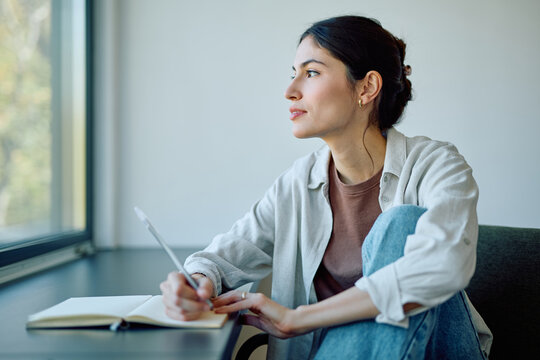 Woman writing thoughts in a journal notebook, pausing to think and look out the window, finding inspiration