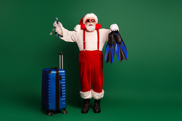 Santa in red costume with beard and hat posing with blue luggage and diving fins for festive...