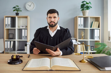 Fototapeta premium Portrait of confident man judge wearing judicial robe looking at camera sitting at the desk with gavel and scales on workplace in courtroom. Male justice working indoors. Legal law concept.