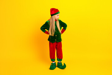Young girl dressed as a festive elf in a bright holiday costume posing against vibrant yellow backdrop celebrating Christmas