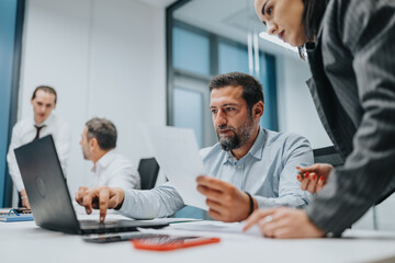 A group of professionals works together in a bright office. A man analyzes a document on paper while a colleague leans over the laptop, sharing ideas during a collaborative business meeting.