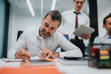 A seasoned manager signs papers at a bright desk while teammates review notes and hold a tablet, capturing collaboration and professional focus in a contemporary office setting.
