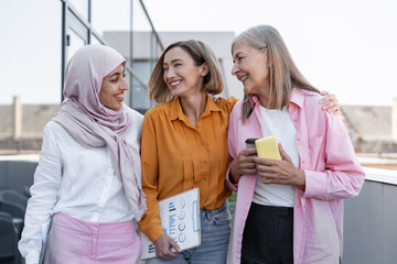 Happy, smiling businesswomen talking, standing together in modern office, one female wearing hijab