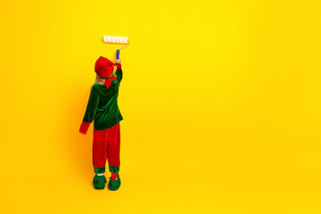 Festive child in vibrant elf costume painting a cheerful wall with a roller for a creative Christmas celebration setting