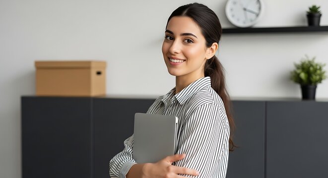 Smiling woman in striped shirt holding laptop in office setting looking at camera