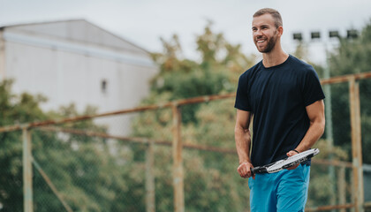 A fit man in a navy shirt and blue shorts stands on an outdoor tennis court, holding a racket and smiling. The scene conveys fitness, sport, and an active, casual lifestyle.
