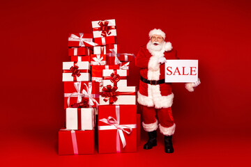 Santa with sale sign beside a tower of gifts in a red studio setting